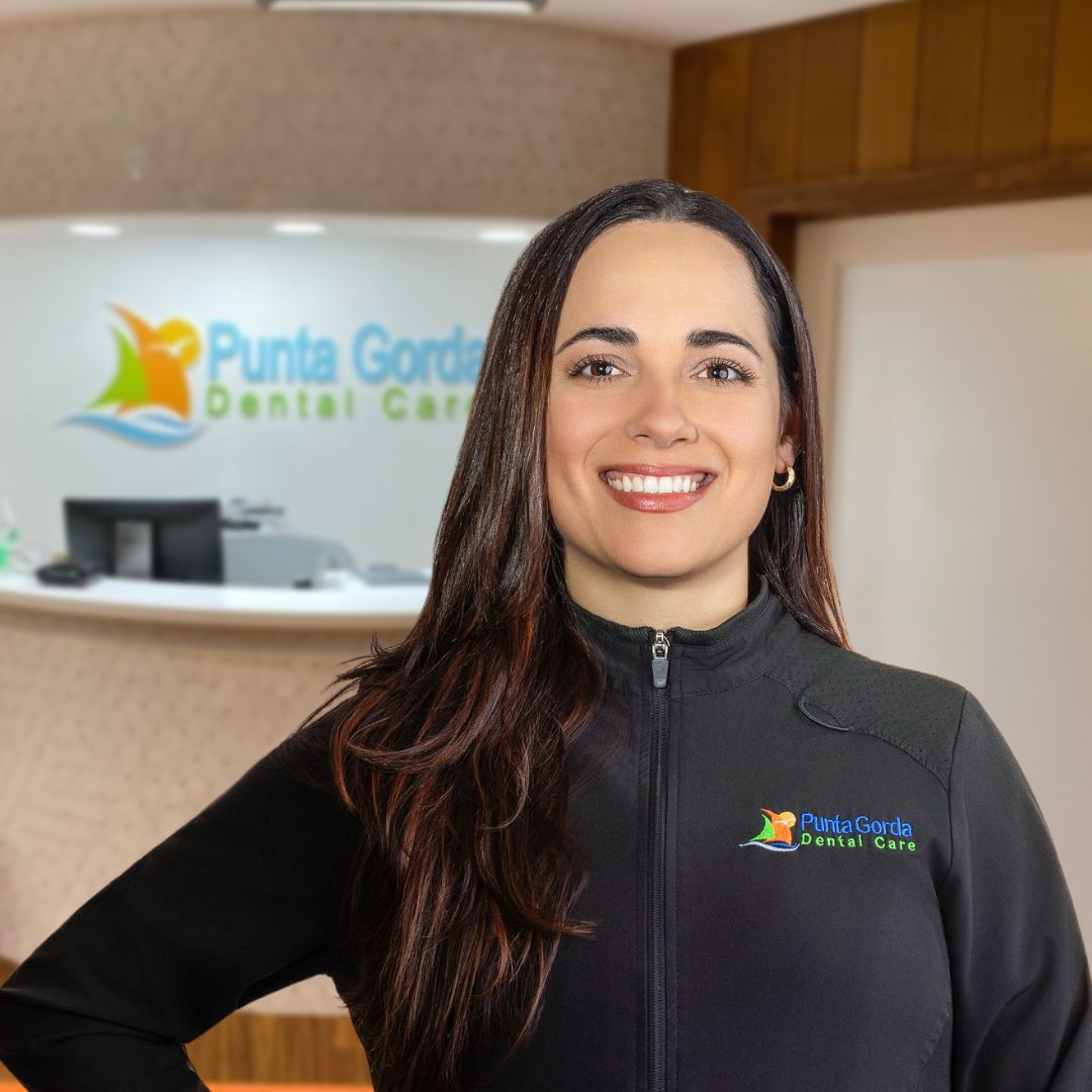 Woman with long dark hair in scrubs jacket smiling and posing in front of Punta Gorda Dental Care front desk, Dr. Marino